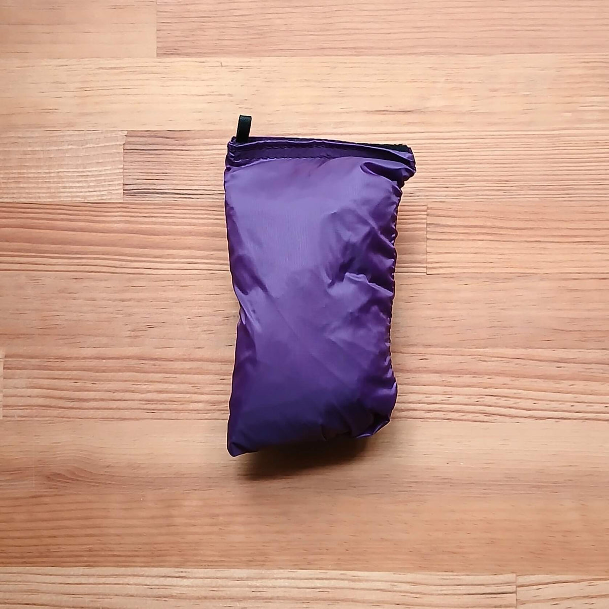 ULA Equipment Rain Kilt レインキルト パープル 一体型収納袋に収めた状態を木目のテーブルに平置きし、袋口中央から立ち上がる黒い引き手タブを写した実物写真 - 山道具とULギアのお店 Ascend Lite