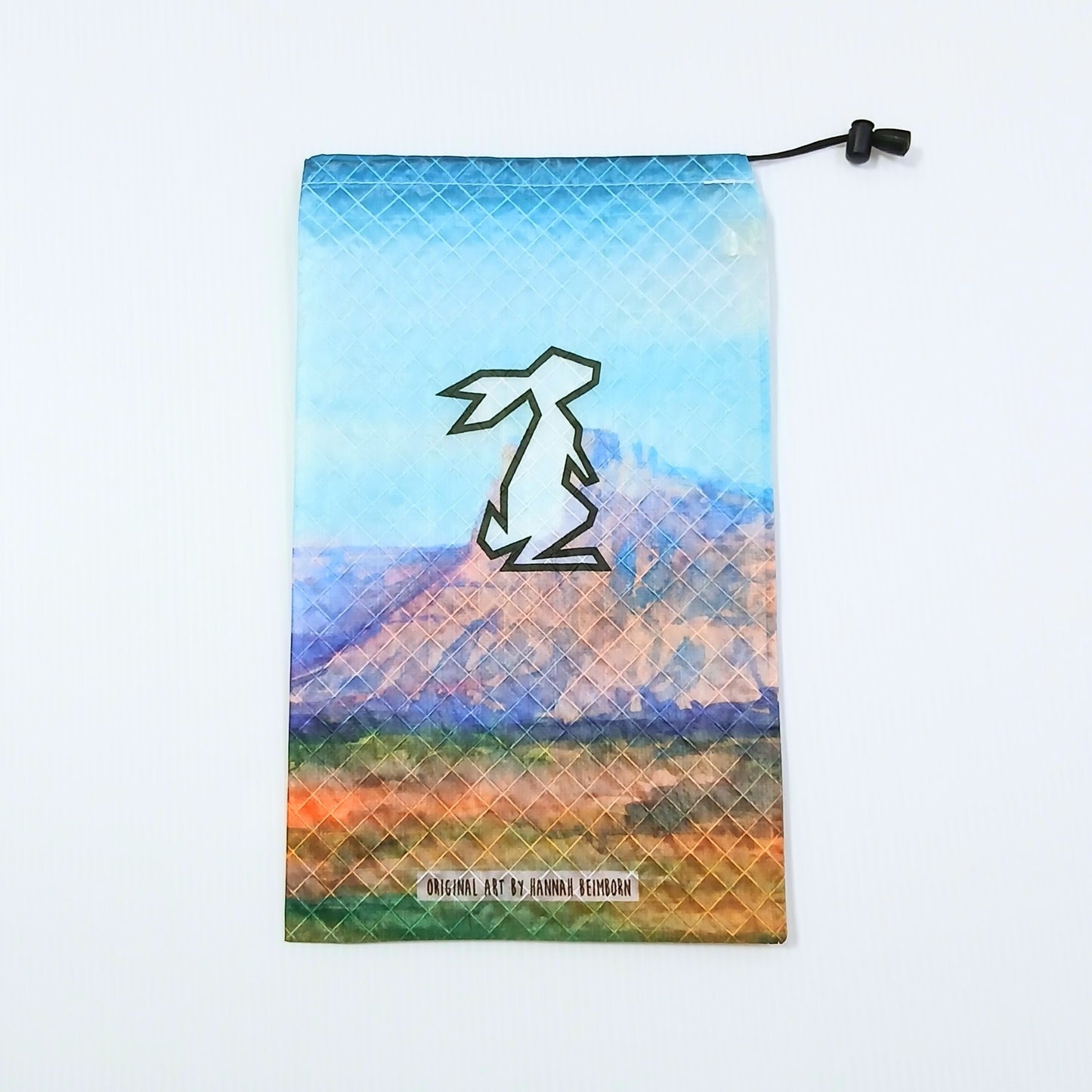 High Tail Designs Ultralight Drawstring Stuff Sack ドローストリングスタッフサック Castle Valley 水色の空とピンク紫のメサを描いた水彩画柄、中央にウサギの輪郭ロゴ、裾にアーティストクレジット印字の実物写真 - 山道具とULギアのお店 Ascend Lite