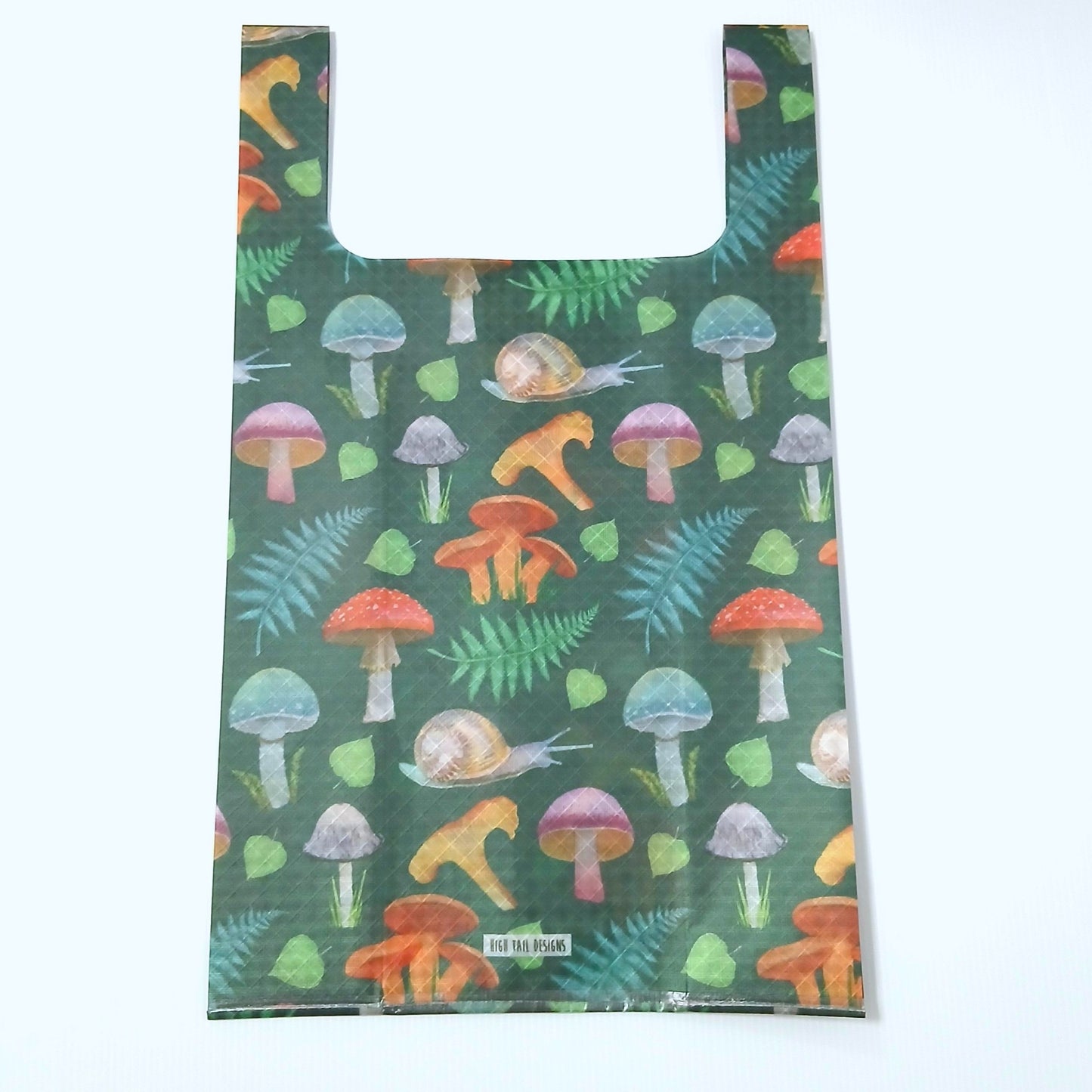 High Tail Designs TX Medium Shopping Bag ショッピングバッグ M Forest Mushroom（Ash Ryan）TX50 同じ深緑のキノコとシダ柄を大きめに裁った個体。細身ハンドルで横幅が絞られた深いシルエットの平置き正面と、下部のアーティストクレジット印字ケアタグの実物写真 - 山道具とULギアのお店 Ascend Lite