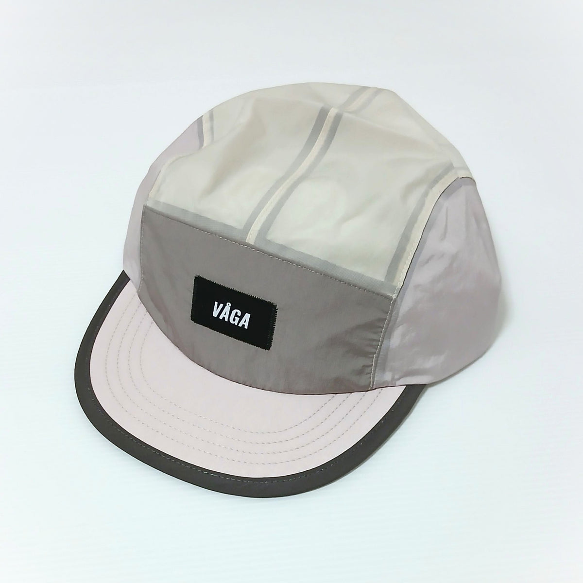 VAGA Pacer Cap ペーサーキャップ ライラック／ダストグレー／スモークグレー 実物写真 斜め前 - スモークグレーのフロントパネル中央の黒いVAGAラベル、生成りのトップパネルと淡いピンクのショートピーク、光沢のあるナイロン生地 - 山道具とULギアのお店 Ascend Lite