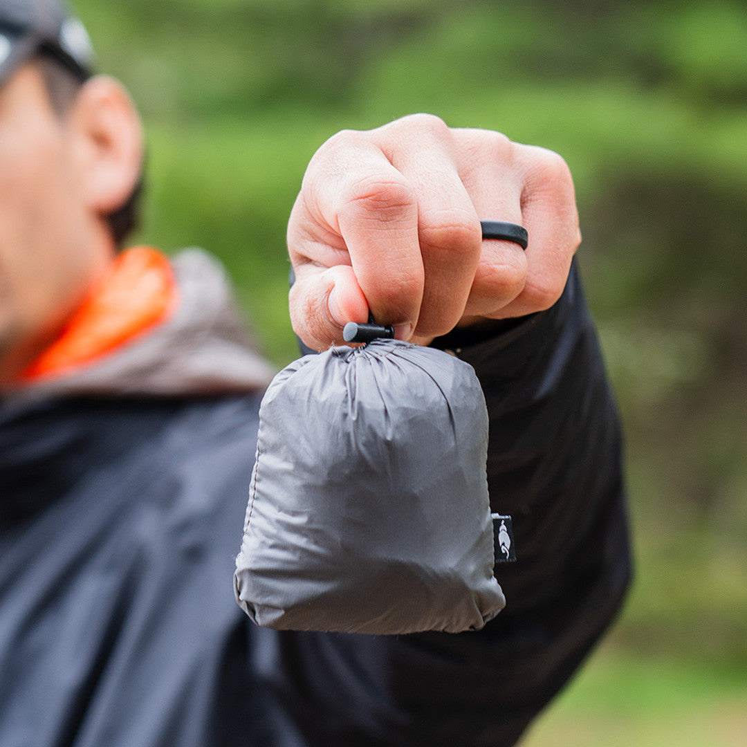 Enlightened Equipment Rain Wrap レインラップ Charcoal 収納した本体を手に持ち握りこぶし大のサイズ感を示した屋外カット（公式画像） - 山道具とULギアのお店 Ascend Lite