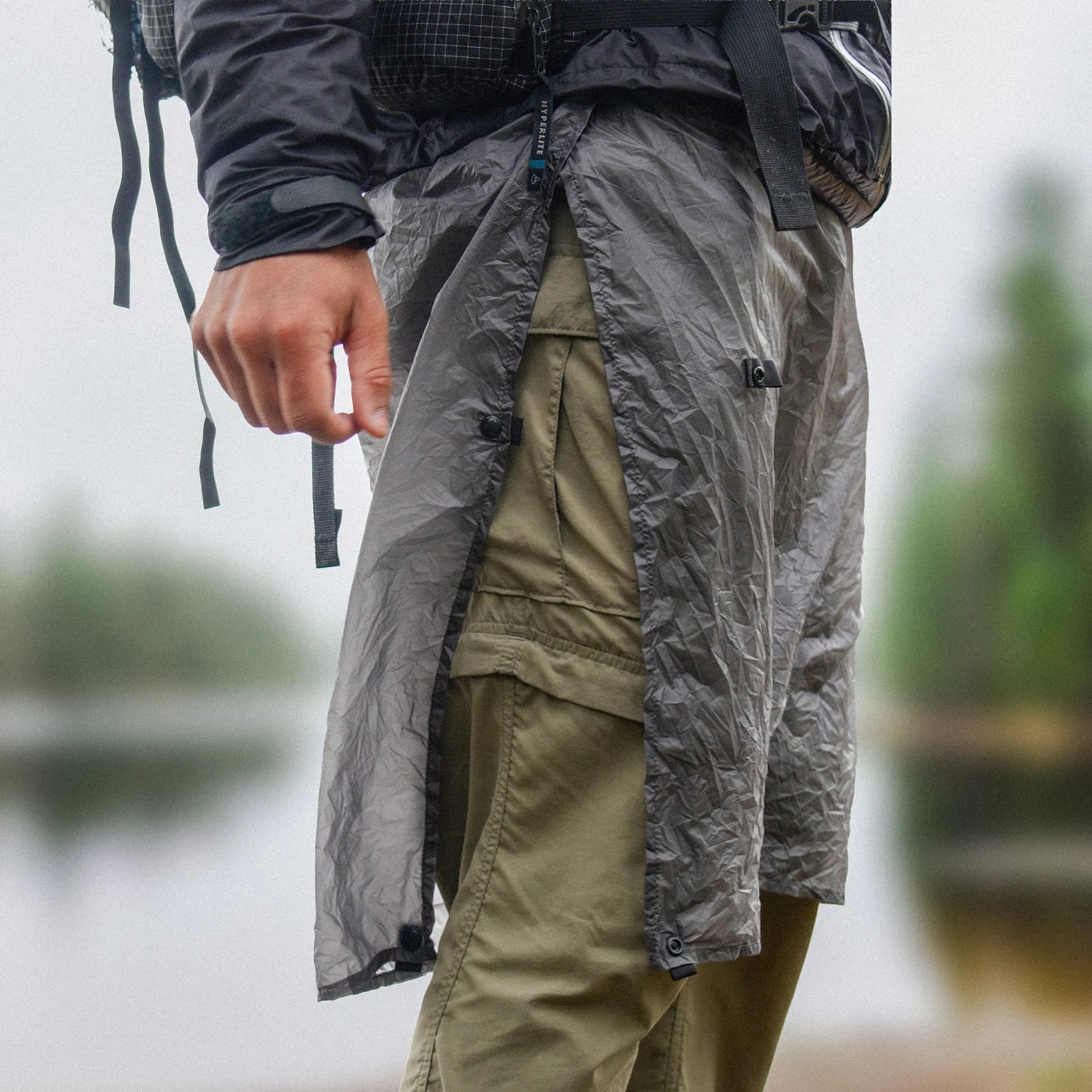 Enlightened Equipment Rain Wrap レインラップ Charcoal 雨の湖畔でザックを背負って歩く着用シーン・合わせが開いて足さばきが確保された状態（公式画像） - 山道具とULギアのお店 Ascend Lite