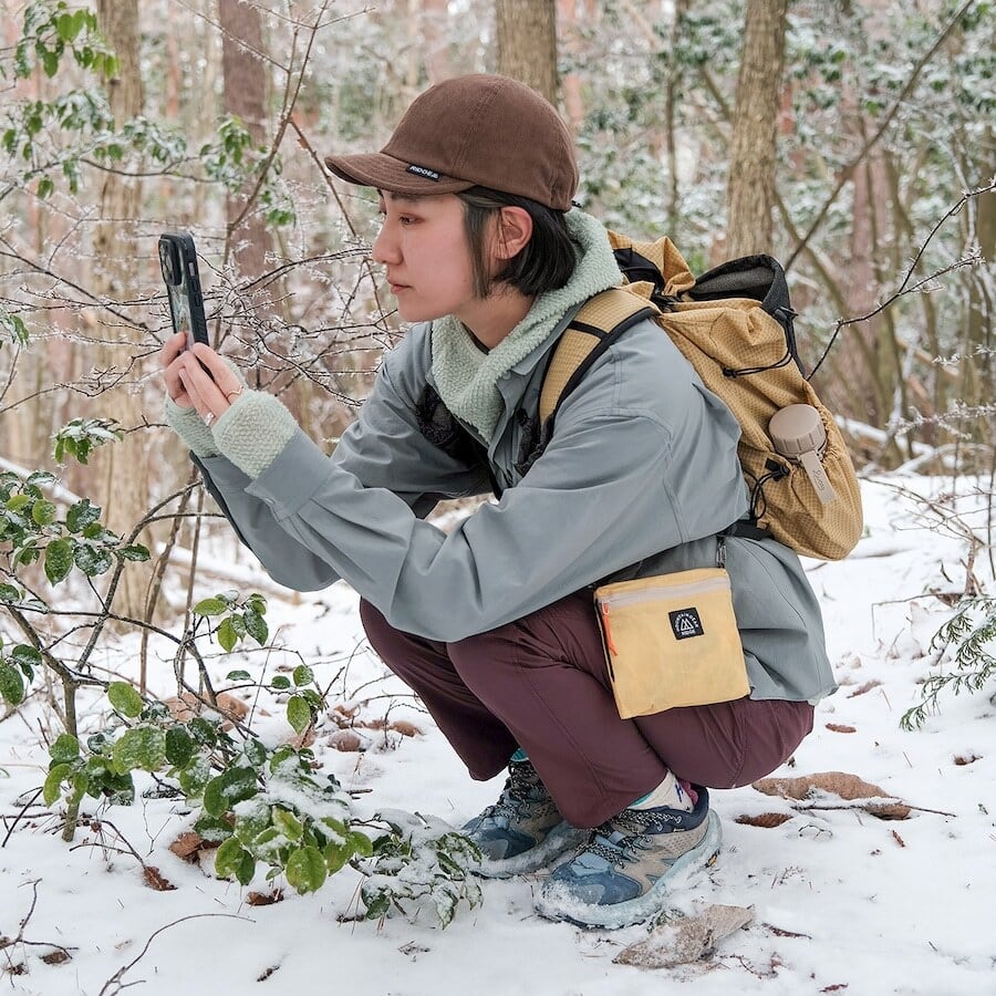 RIDGE MOUNTAIN GEAR Travel Pouch Plus トラベルポーチプラス Wheat Yellow を腰に付け雪の森でしゃがんでスマートフォンを使う使用シーン（公式画像） - 山道具とULギアのお店 Ascend Lite