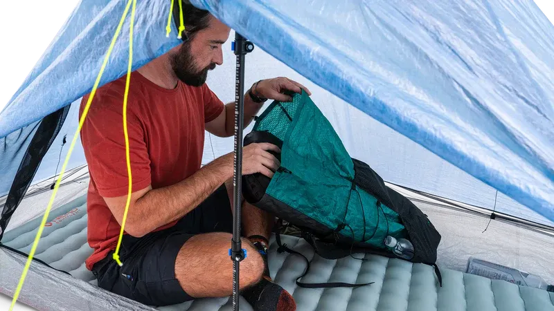 Zpacks Plex Solo Pro Tent プレックスソロプロテント Blue 室内 - 入口脇に座り、中央のトレッキングポール越しにバックパックを室内へ引き込む様子と、光を透かす半透明のDCFキャノピー（公式画像） - 山道具とULギアのお店 Ascend Lite